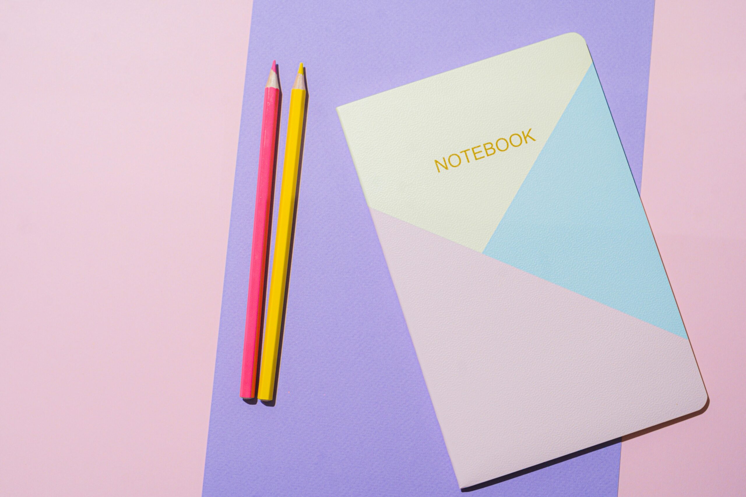 A minimalist flat lay of a geometric notebook and colored pencils on pastel backgrounds.